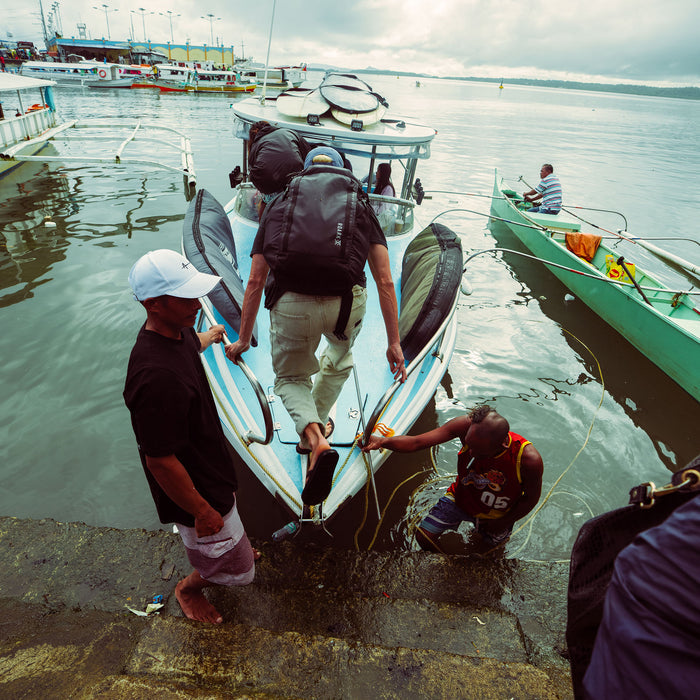 Hiker wearing black Accomplice Mule 45L backpack while loading boat at coastal dock with water and vessels visible