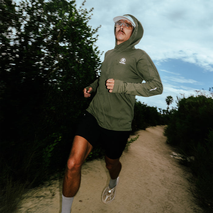 Man wearing light military green Alta Elite Trail Hood while jogging on a dirt path surrounded by green vegetation and trees