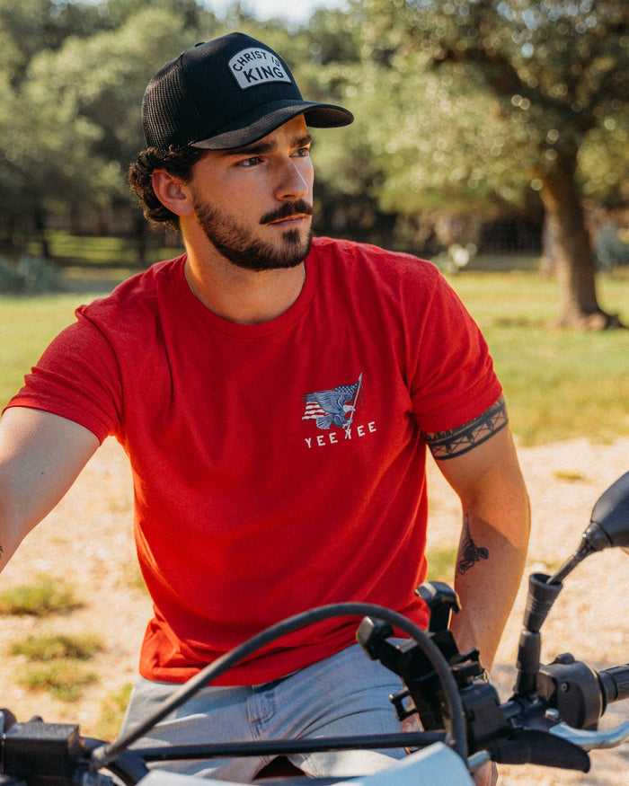 Man wearing a red t-shirt with a logo and a black cap, sitting on a motorcycle in a natural setting.