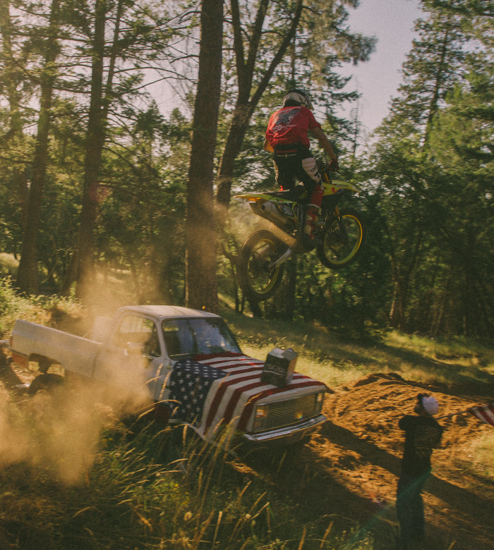 Person on a dirt bike performing a jump over an old car with an American flag, in a forest setting.