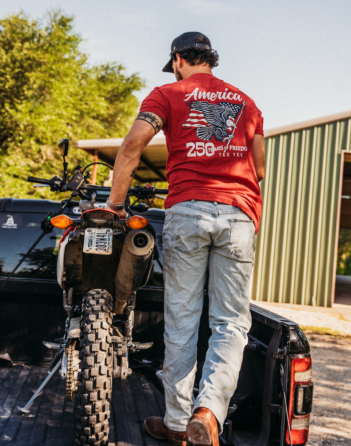 Man in a red shirt with 'America' and '250' design standing next to a motorcycle in a truck bed.