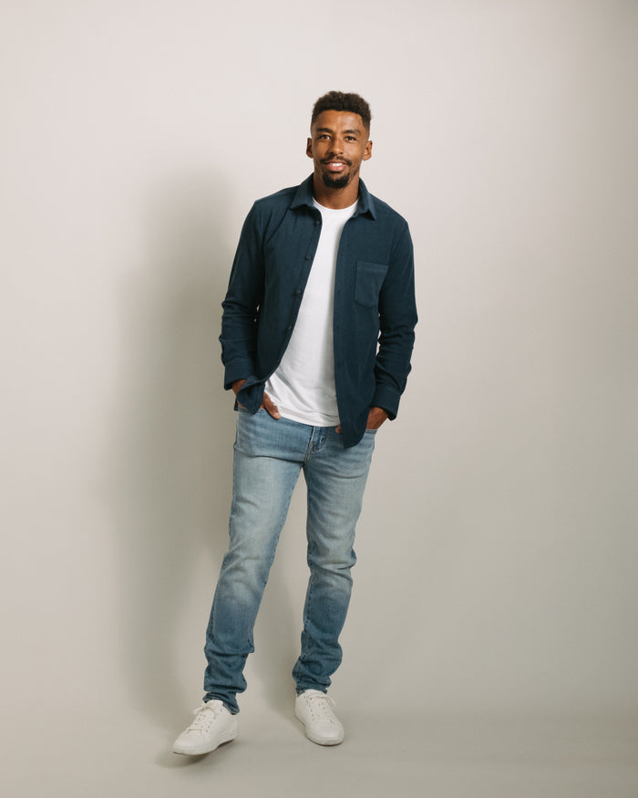 Man wearing a storm blue flannel button-up shirt, white t-shirt, blue jeans, and white sneakers on a plain background