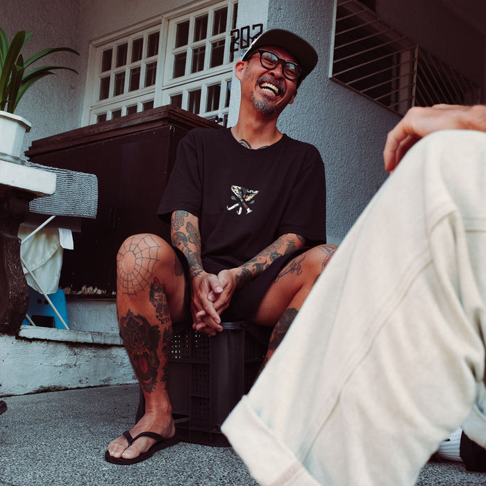 Man wearing black Augs Safe Camp Premium Tee with small chest graphic, seated outdoors in lifestyle shot showing shirt fit and design detail