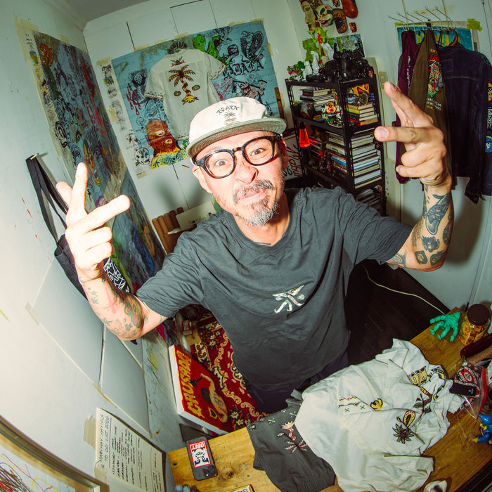 Man wearing washed black short-sleeve tee with small logo, posing in art studio surrounded by colorful artwork and clothing