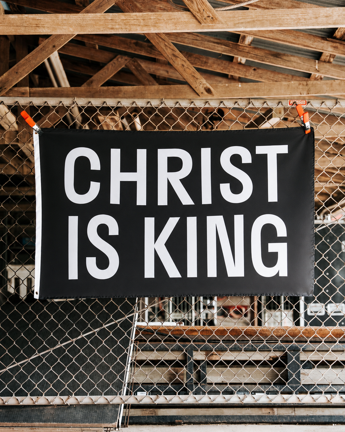 Black banner with 'CHRIST IS KING' text hanging against a wooden and wire fence background