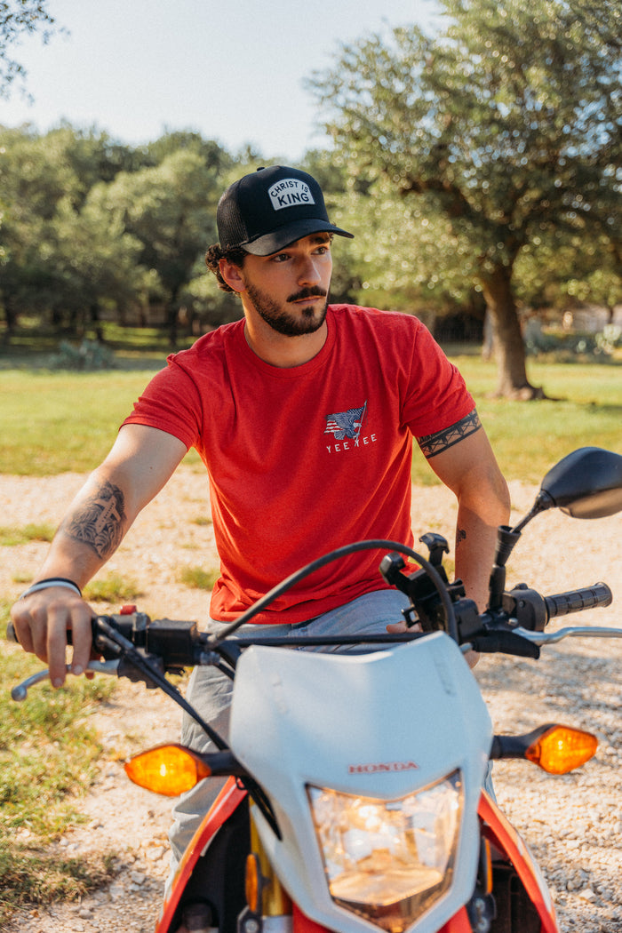Man in a red shirt and black cap sitting on a Honda motorcycle in a natural setting.