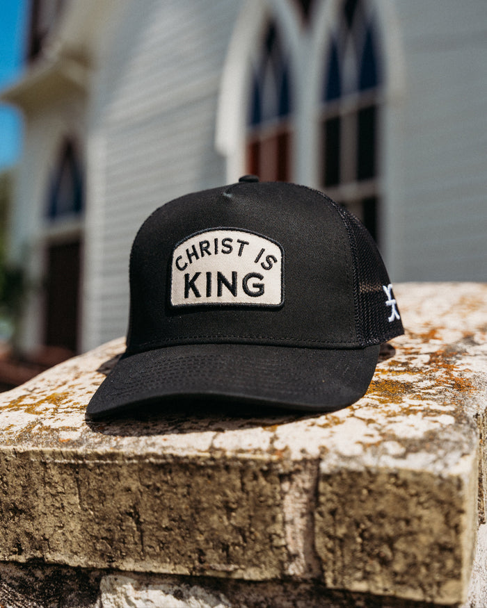 Black cap with 'Christ is King' patch on a stone ledge with a blurred church in the background