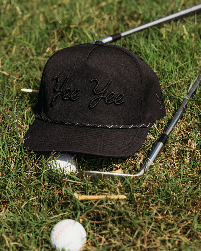 Black cap with 'Yee Yee' text on grass with golf club and ball