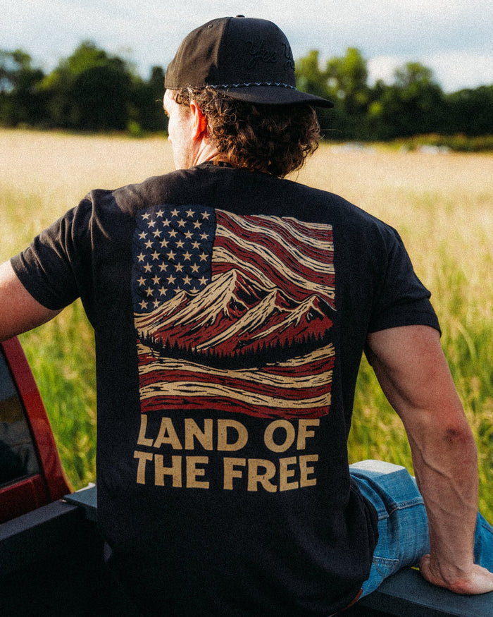 Person wearing a t-shirt with an American flag design and 'Land of the Free' text, sitting in a field.