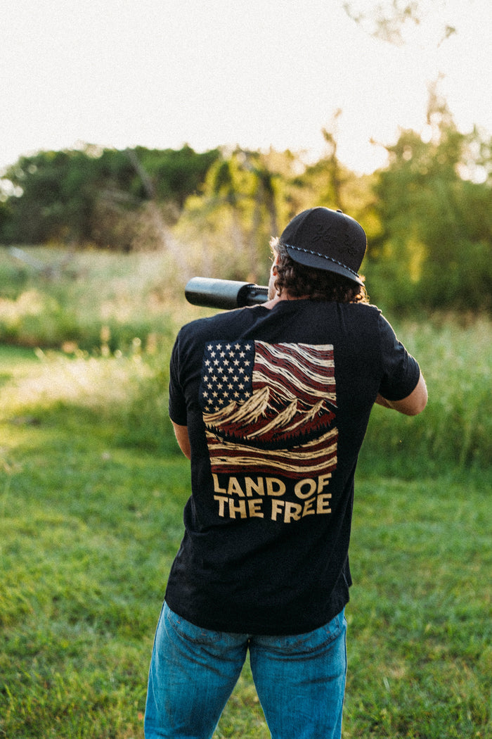 Person wearing a black t-shirt with an American flag design and 'Land of the Free' text, standing in a grassy field.