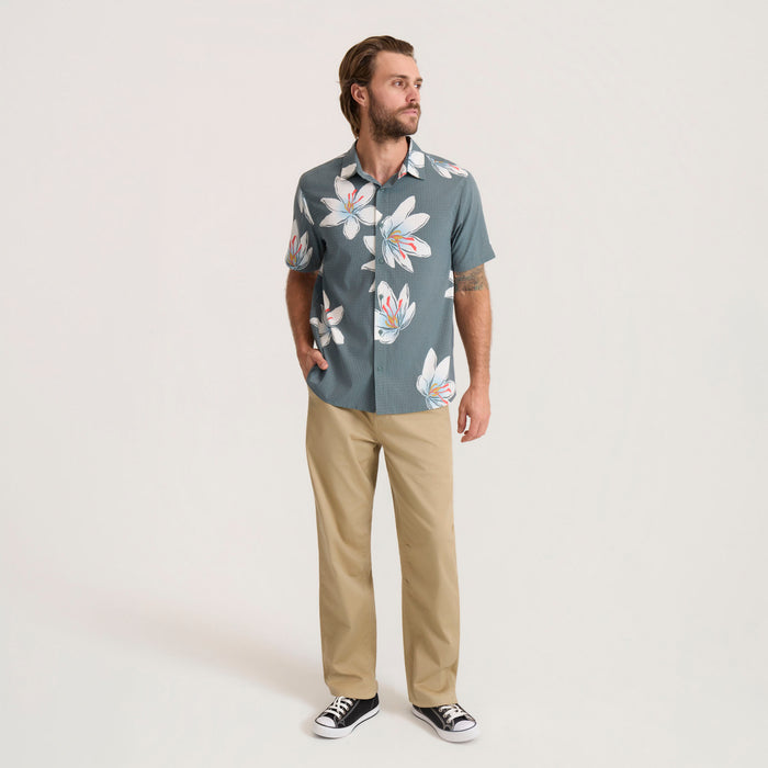 A man wearing a slate blue short-sleeve button-down shirt with a white and pink hibiscus floral print, paired with khaki pants and black sneakers, standing against a light gray studio background.