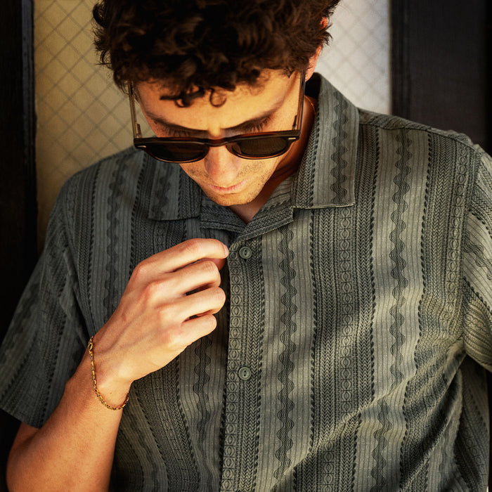 Man with dark curly hair wearing gray patterned short-sleeve shirt and dark sunglasses, looking downward