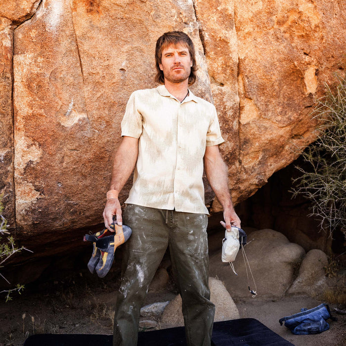 Man wearing sand-colored short-sleeve button-up shirt standing against weathered terracotta stone wall holding climbing gear