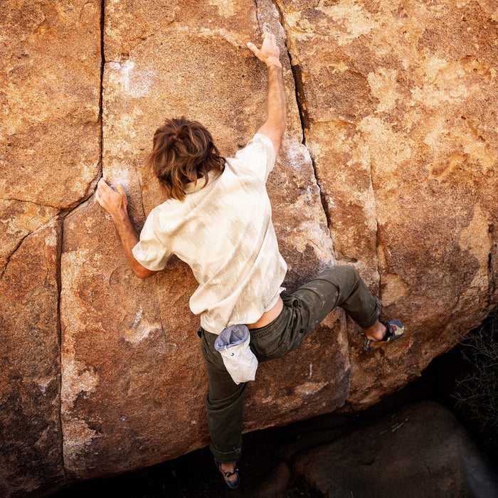 Man rock climbing wearing sand-colored Bless Up Trail Button Up Shirt with dark pants and white climbing gloves on textured rock face