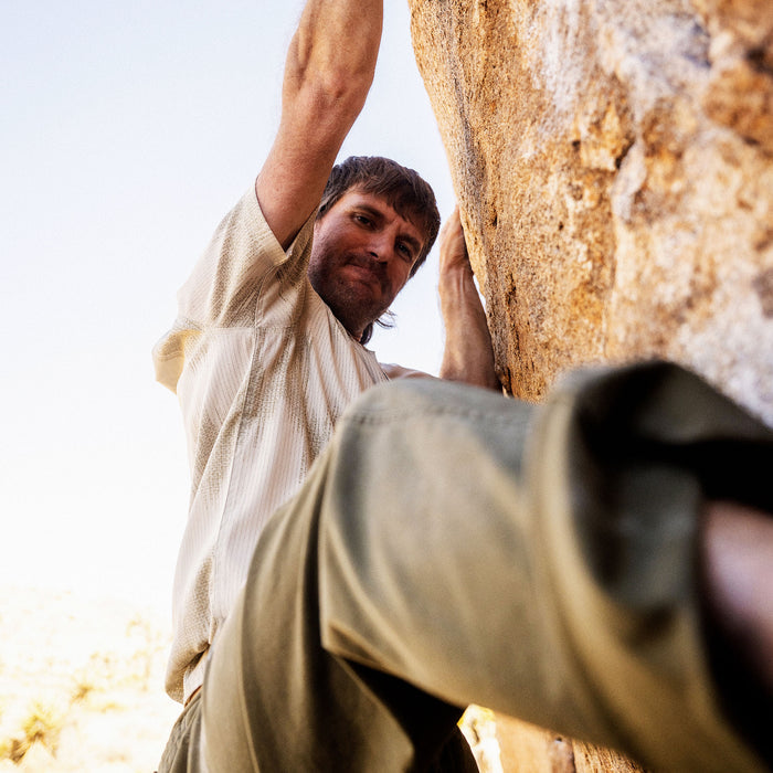 Man wearing sand-colored Bless Up Trail Button Up Shirt climbing textured rock wall in outdoor lifestyle photo