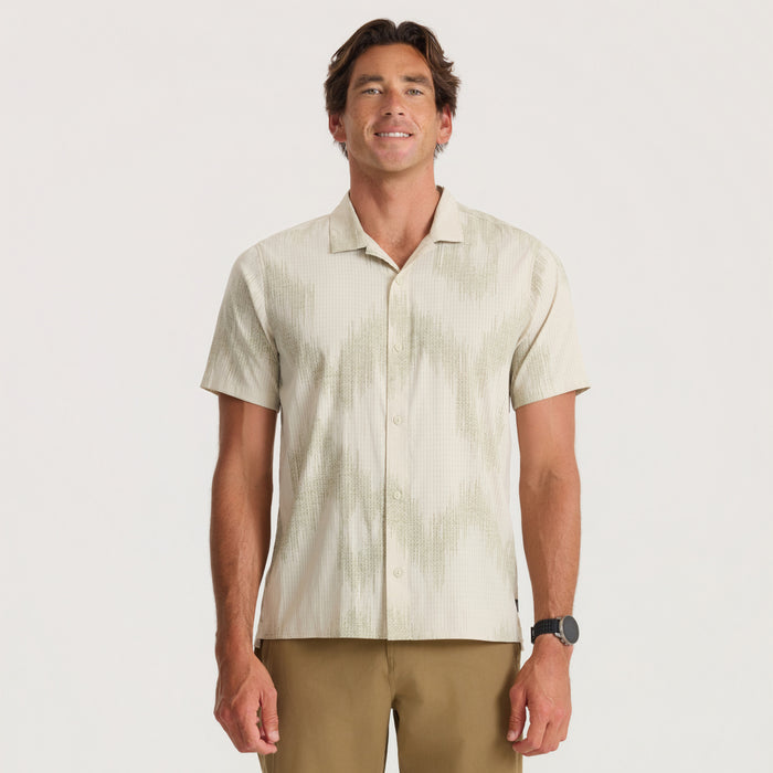 Man wearing cream textured short-sleeve button-up shirt with sand-colored pants, standing against neutral background