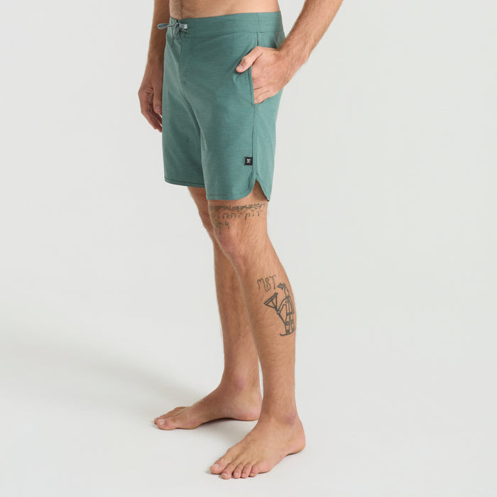 Surfer clad in Roark boardshorts carrying a surfboard on a sunny beach, embodying the spirit of adventure and freedom.