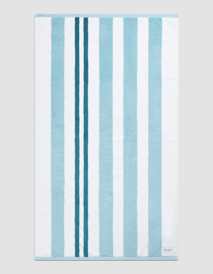 The Classic Stripe Resort Towel by Cozy Earth in Wave Stripe is shown laid flat on a light gray background. Alternating vertical blue and white stripes and a small white label in the bottom right corner are visible.|Color:Wave Stripe