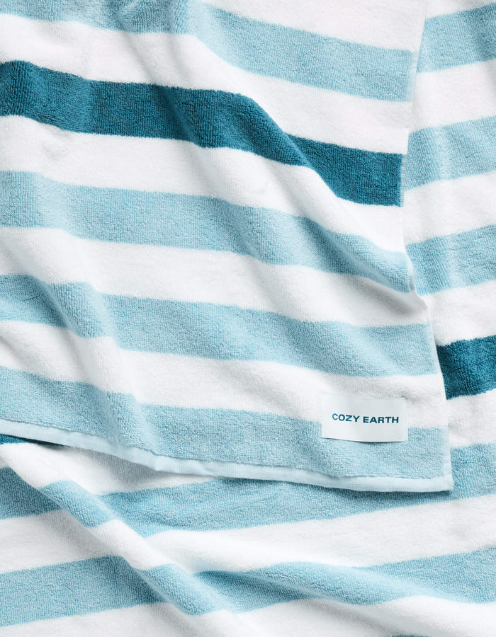 The Classic Stripe Resort Towel by Cozy Earth is a soft towel with wide blue and white horizontal stripes, featuring a small label that reads "Cozy Earth.|Color:Lake Stripe