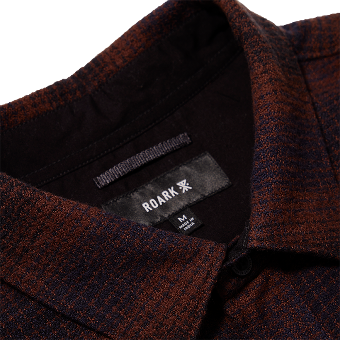 Close-up of a high-quality woven fabric with detailed woolen texture, featuring a button, label, and pocket on a Roark adventure-inspired garment.