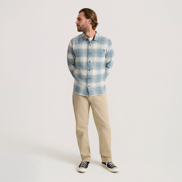 Man wearing blue and cream plaid flannel shirt with beige chinos and dark sneakers, standing against neutral background