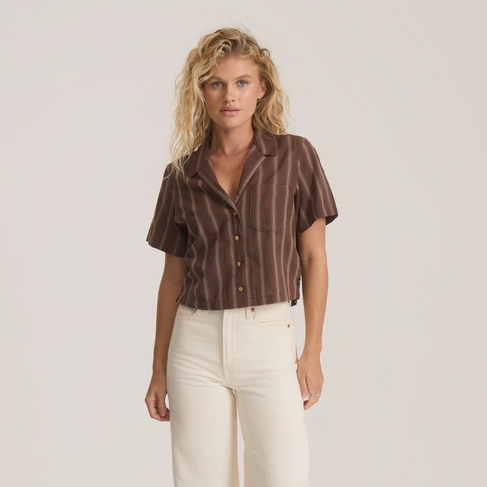 Brown striped camp button up shirt with short sleeves styled on model wearing cream pants against neutral background