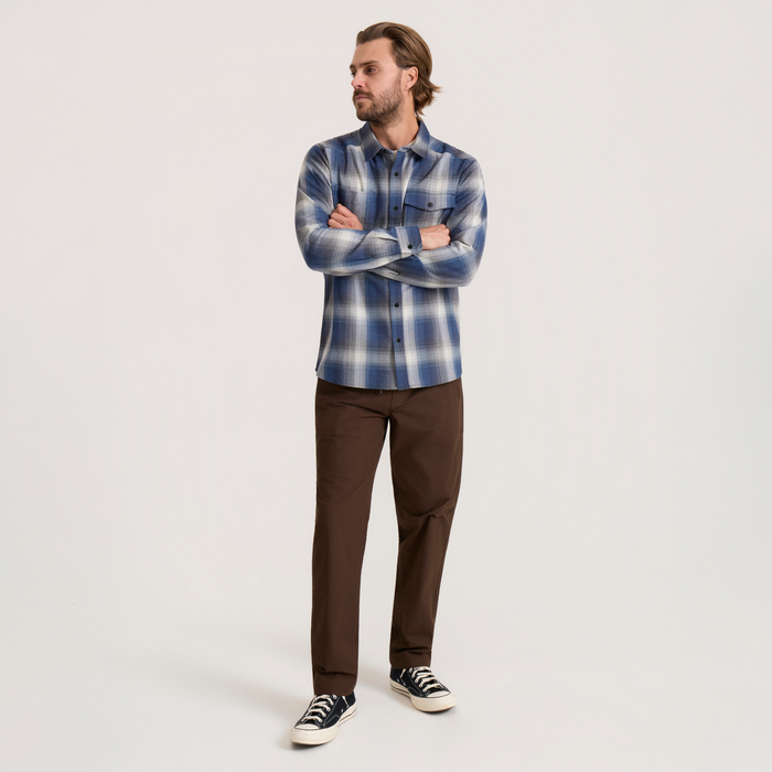 Man wearing slate blue and white plaid flannel shirt with brown pants and black canvas sneakers, standing with arms crossed