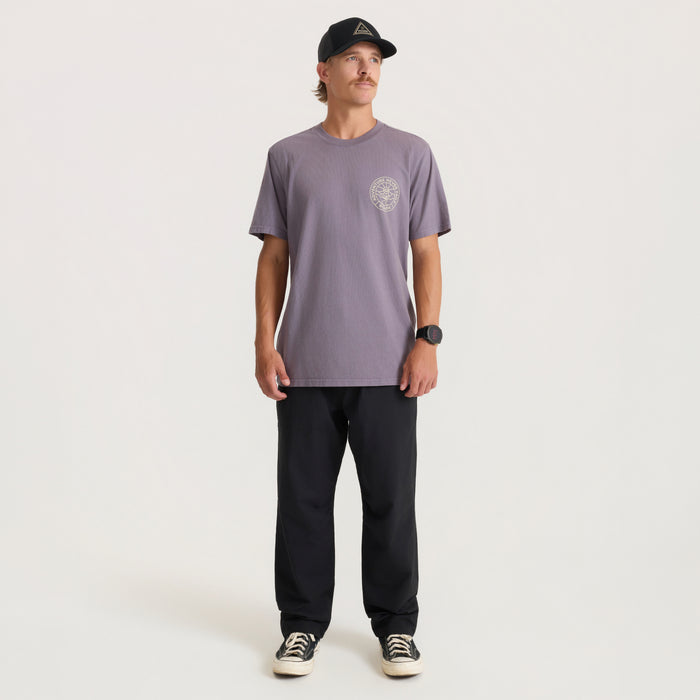 Man wearing mauve Division Premium Tee with chest graphic, black pants, and black cap, standing against neutral background