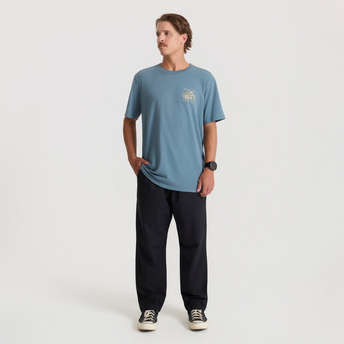 Slate blue short sleeve tee with small chest graphic worn by model with black pants and canvas sneakers, full body shot