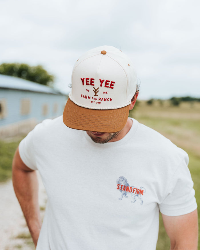 Man wearing a white cap with 'Yee Yee' text and a white t-shirt with a logo, standing outdoors.