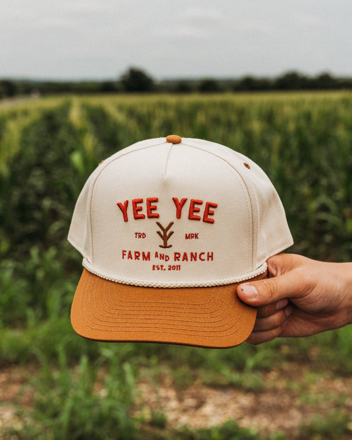 Cap with 'Yee Yee Farm and Ranch' text held in front of a cornfield
