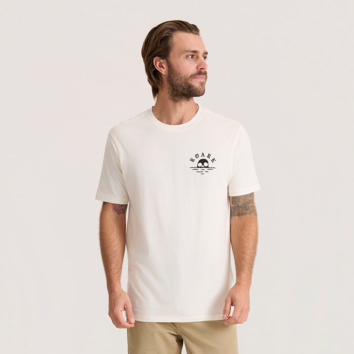 Men's cream short-sleeve tee with small black skull and wave graphic on chest, worn with khaki pants
