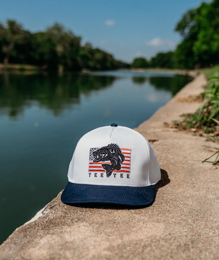 White cap with blue brim featuring a fish design on a concrete ledge by a lake.