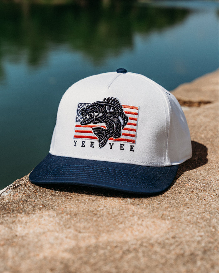 White cap with black brim featuring a fish graphic and 'Yee Yee' text on a concrete ledge by a body of water.