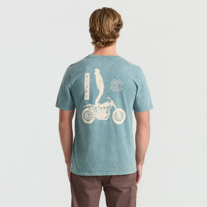 Active cyclist in motion wearing Roark adventure shirt with a detailed bicycle frame in a natural setting.