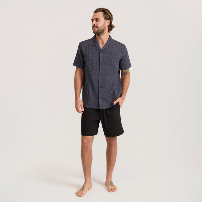 Man wearing charcoal gray short-sleeve button-up shirt with vertical striped pattern and black shorts in full-body lifestyle shot