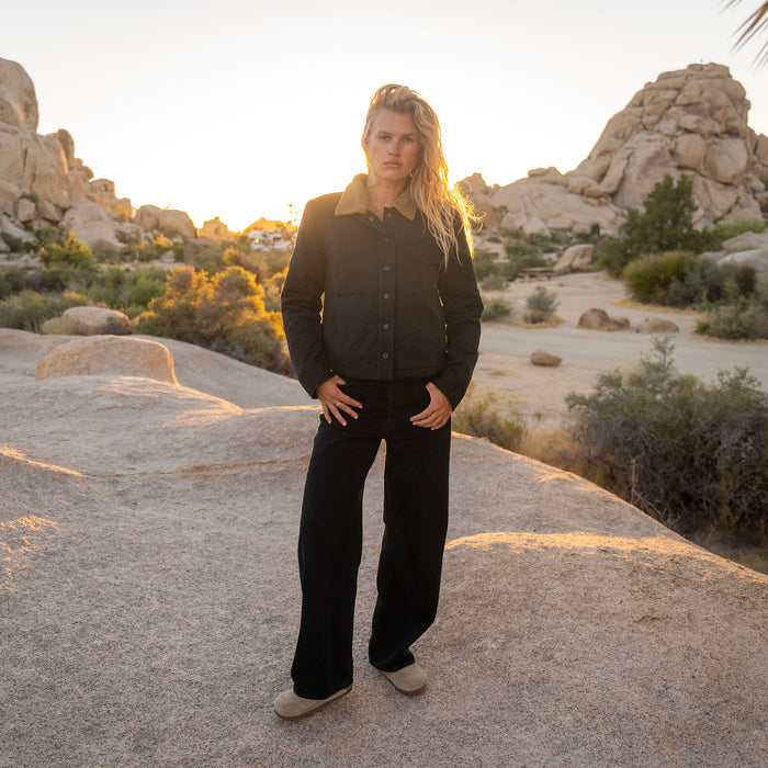 Woman wearing black Hebrides Sherpa jacket with sherpa collar and black pants, standing in desert landscape at sunset