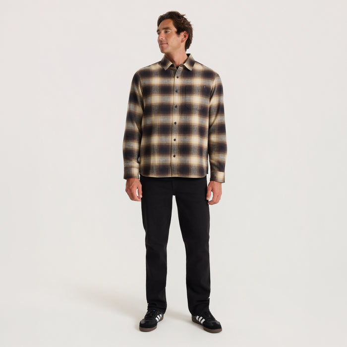 Man wearing brown and cream plaid button-up shirt with black denim jeans and black sneakers, standing against white background