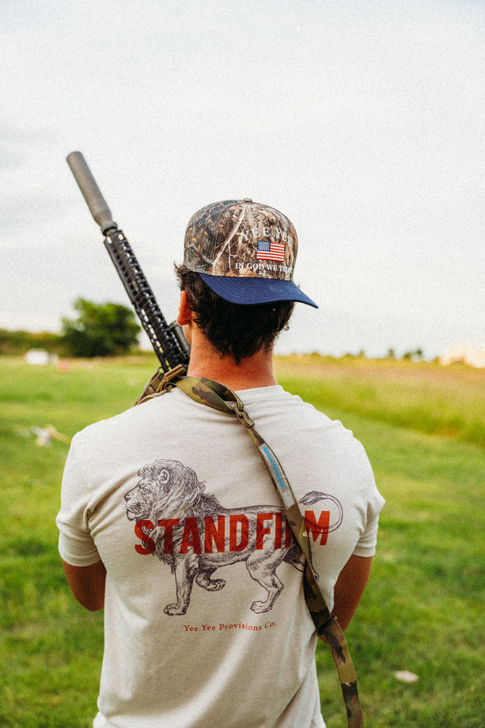 Man with rifle on shoulder wearing a cap and 'Stand Firm' t-shirt in a field