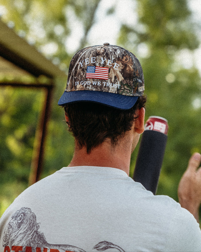Person wearing a camouflage cap with text and an American flag, standing outdoors with trees in the background.