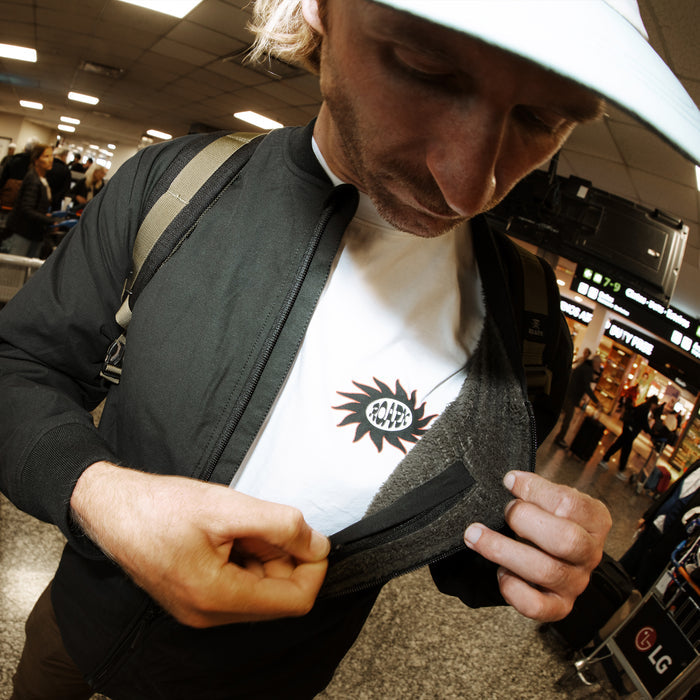 Man wearing black Layover Bomber Jacket with white undershirt, showing interior pocket detail in airport setting