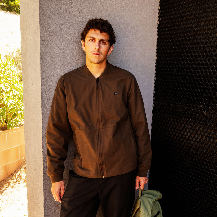 Man wearing tobacco brown Layover Bomber Jacket with black pants, standing against gray wall in outdoor setting