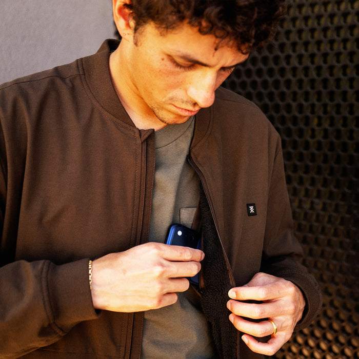 Man wearing tobacco brown Layover Bomber Jacket over gray shirt, holding phone in jacket pocket during lifestyle shot