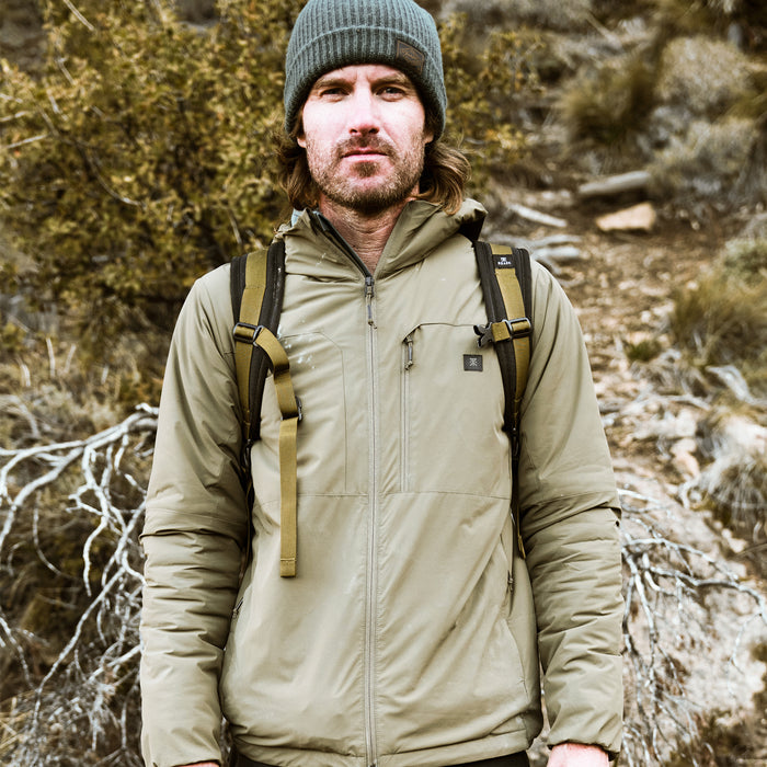 Man wearing gray beanie and tan insulated Layover Jacket with tan backpack straps, standing outdoors among evergreen trees