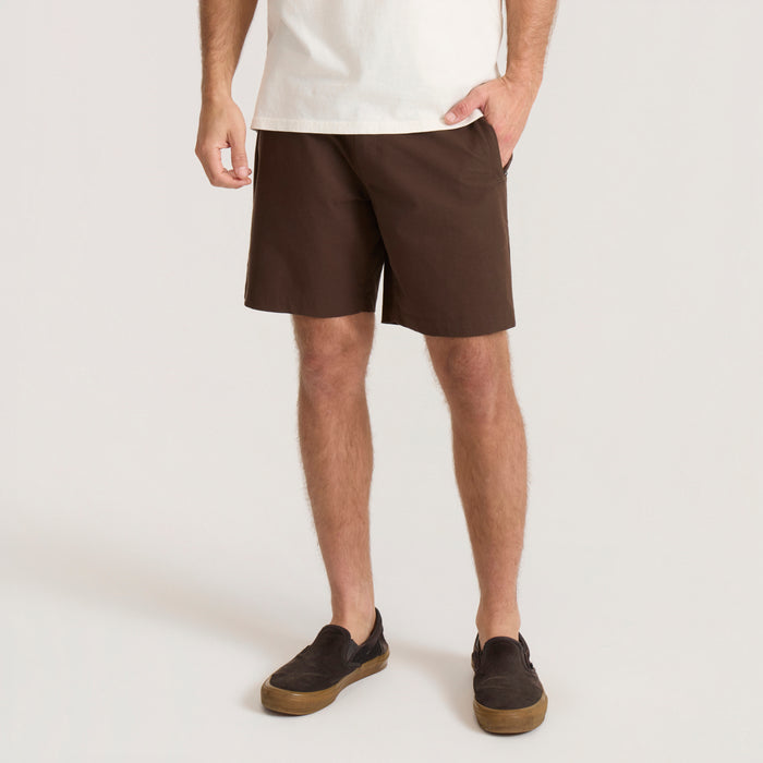 Man wearing tobacco brown Layover Pro 8" Traveler Shorts with white t-shirt and black slip-on shoes on neutral background