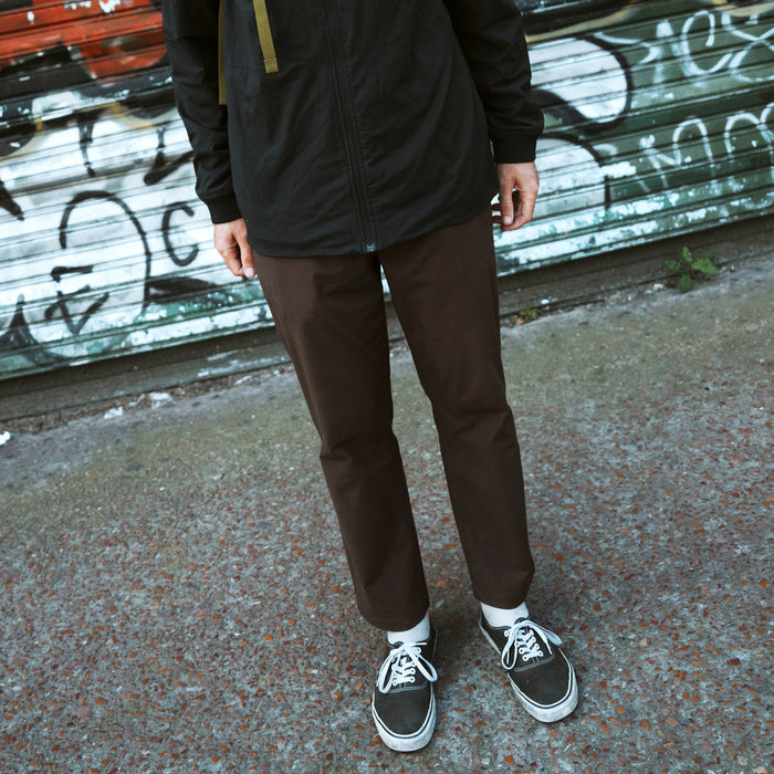 Person wearing tobacco brown Layover Pro Traveler Pants with black jacket and black and white sneakers, standing outdoors