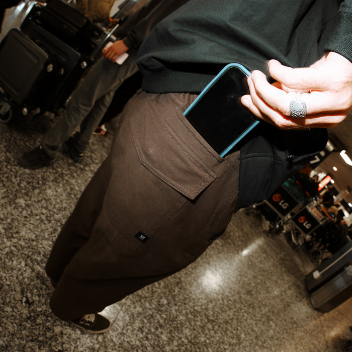 Person wearing tobacco brown Layover Pro Traveler Pants with cargo pocket, holding blue smartphone, lifestyle shot