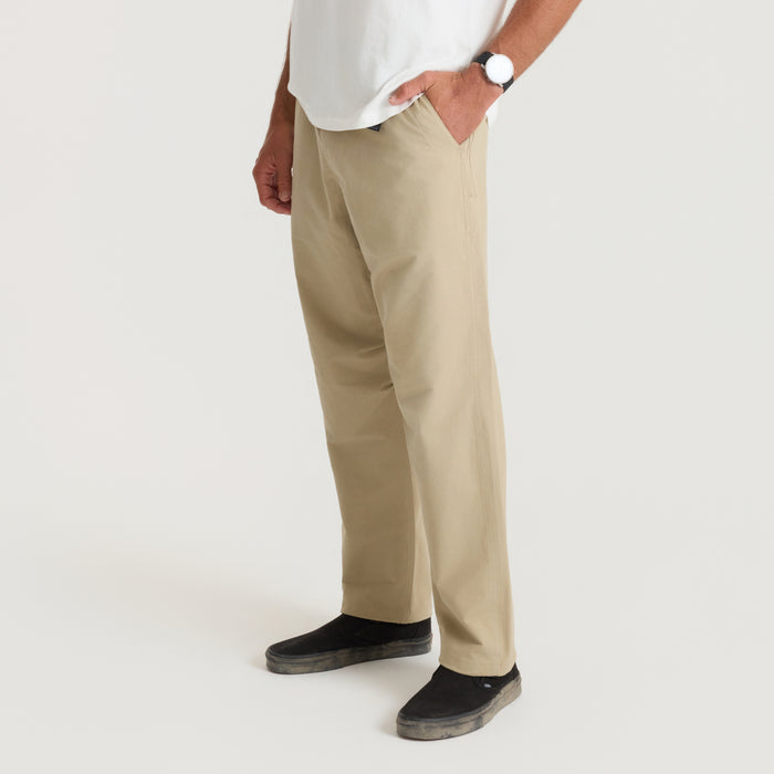 Beige Layover Trail Pants worn by man on model with white shirt and black slippers, side view showing relaxed fit and full length
