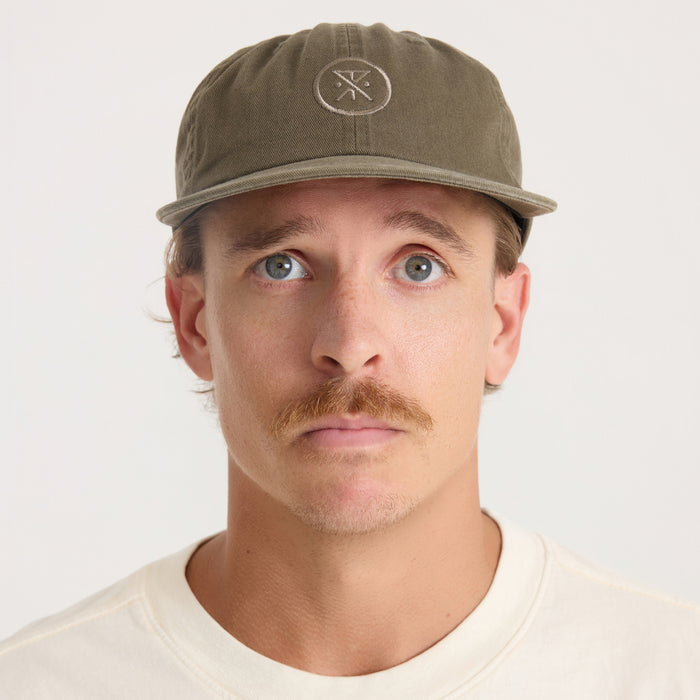 Washed military green six-panel strapback hat with embroidered circular logo, worn on model with cream t-shirt against neutral background