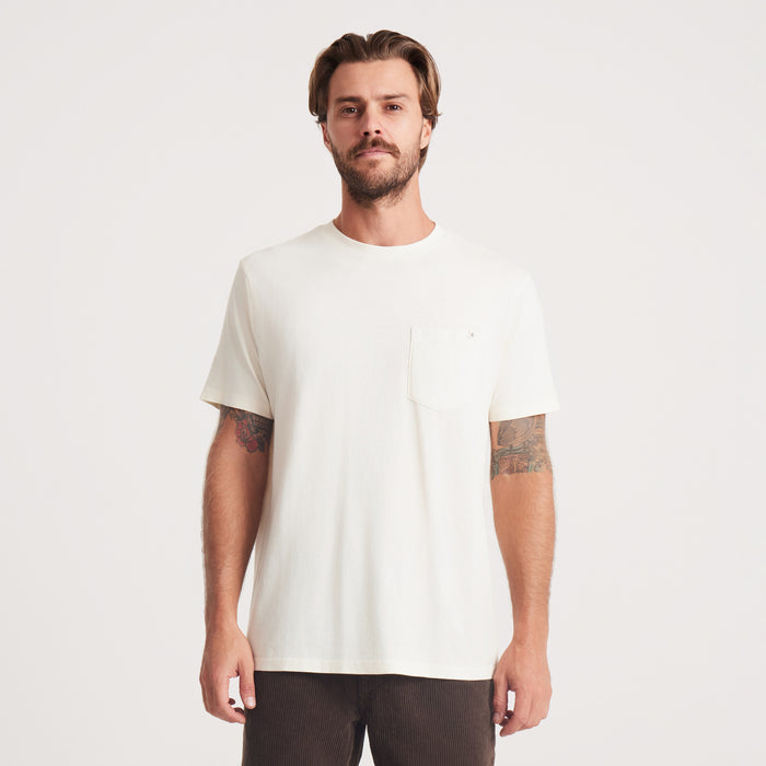 A man stands confidently wearing a white Roark t-shirt, featuring a pocket on the left side, showcasing casual style and comfort.